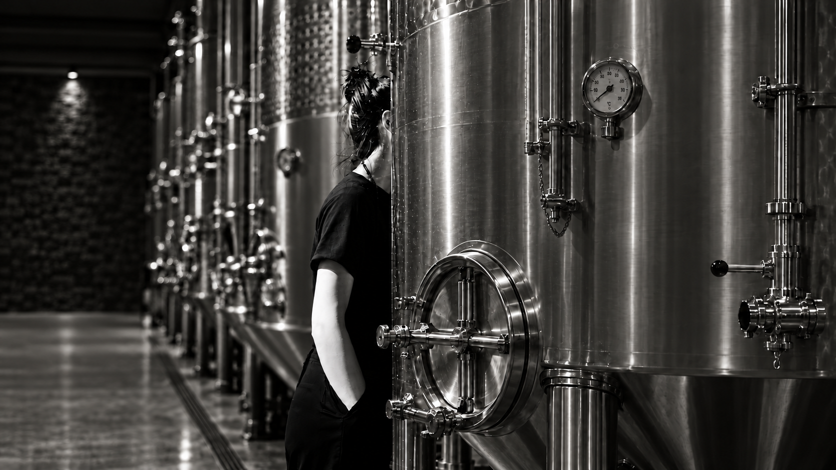 A person among stainless-steel fermentation tanks in a winery, photographed in black and white